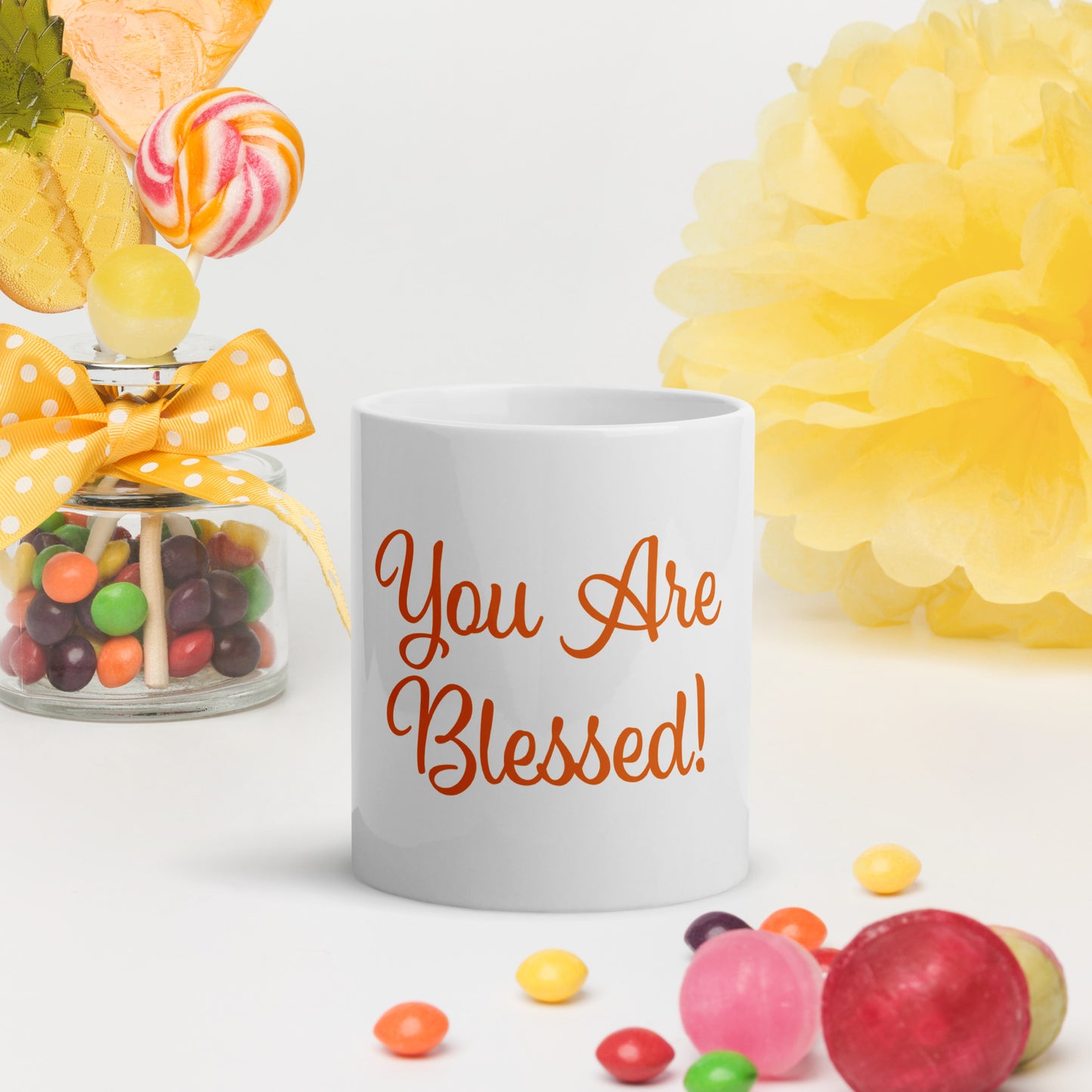 White mug with You Are Blessed text, surrounded by colorful candies and flowers on a white background.